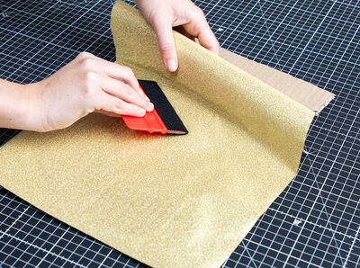 Rakel streicht Klebefolie Glitter gold auf Kartonziffer fest.