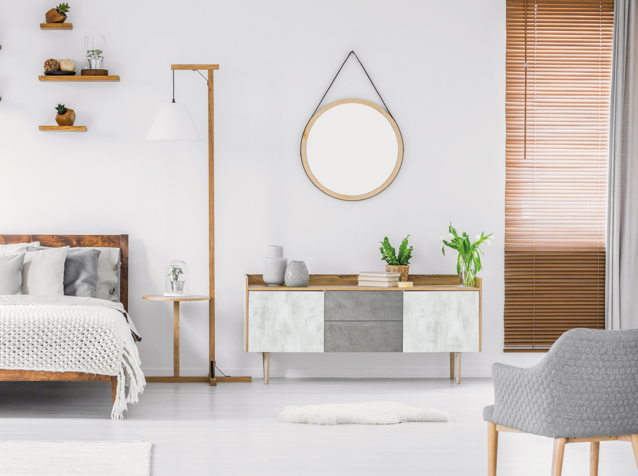 Modern eingerichtetes Schlafzimmer mit M&ouml;beln in Holzoptik, heller Kommode, rundem Wandspiegel und braunen Jalousien.