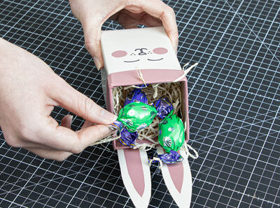 Hand legt bunte Schokoeier auf das Papiergras im Osternest. 