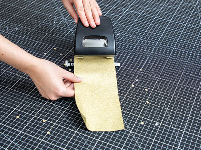 Neben Locher liegt Konfetti aus goldfarbener Metallic-Glitterfolie. 