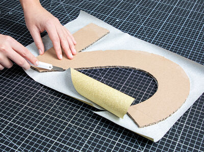 Ziffern liegen mit der Kartonseite nach oben, Hand mit Cutter schneidet an Ziffer &uuml;berstehende Klebefolie ab.